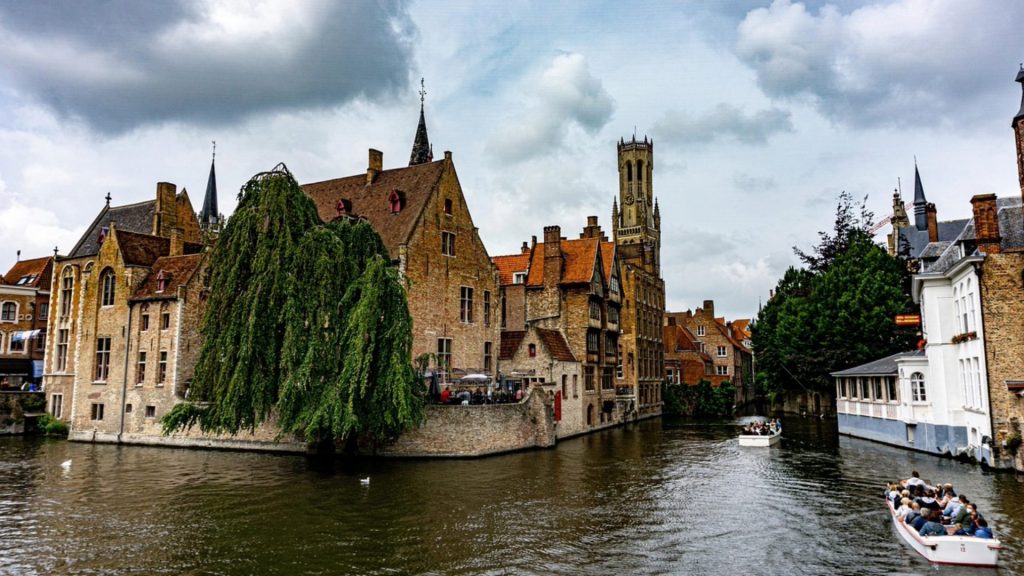 A picturesque canal scene in Bruges with tourist boats gliding past medieval buildings and a weeping willow, capturing the city's nickname as "Venice of the North."