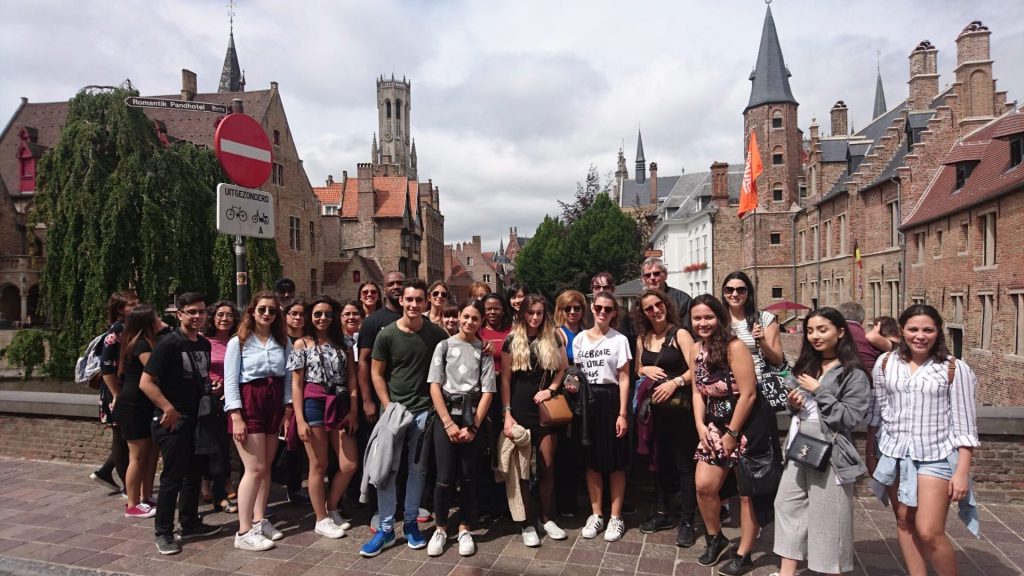 A student tour group posing for a photo in front of Bruges' medieval architecture, with the famous Belfry tower visible in the background.