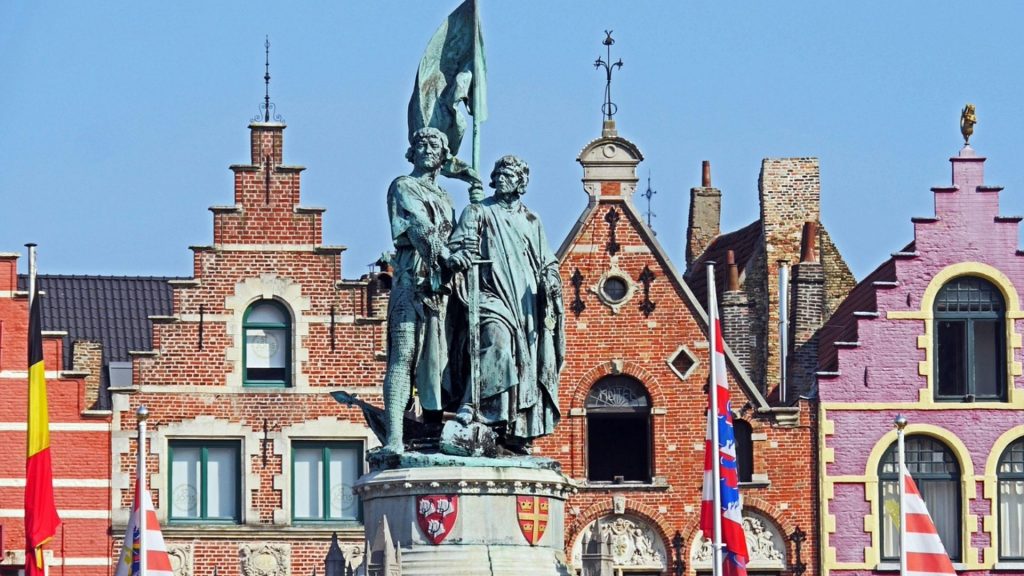 The Jan Breydel and Pieter de Coninck statue in Bruges' central square, surrounded by colorful traditional stepped-gable buildings characteristic of Flemish architecture.
