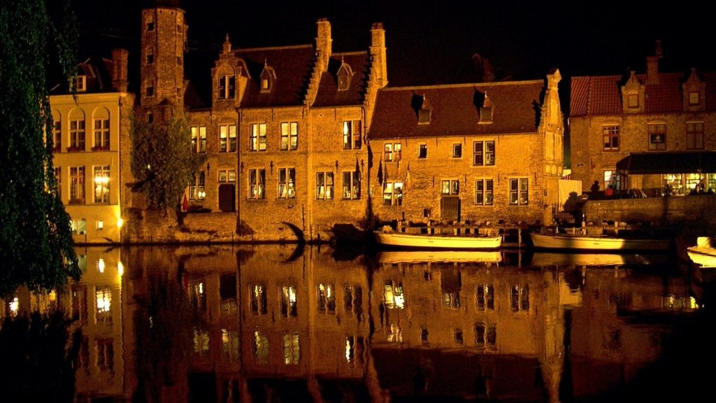 Medieval buildings illuminated at night along the canal in Bruges, their warm glow reflecting perfectly in the still waters.
